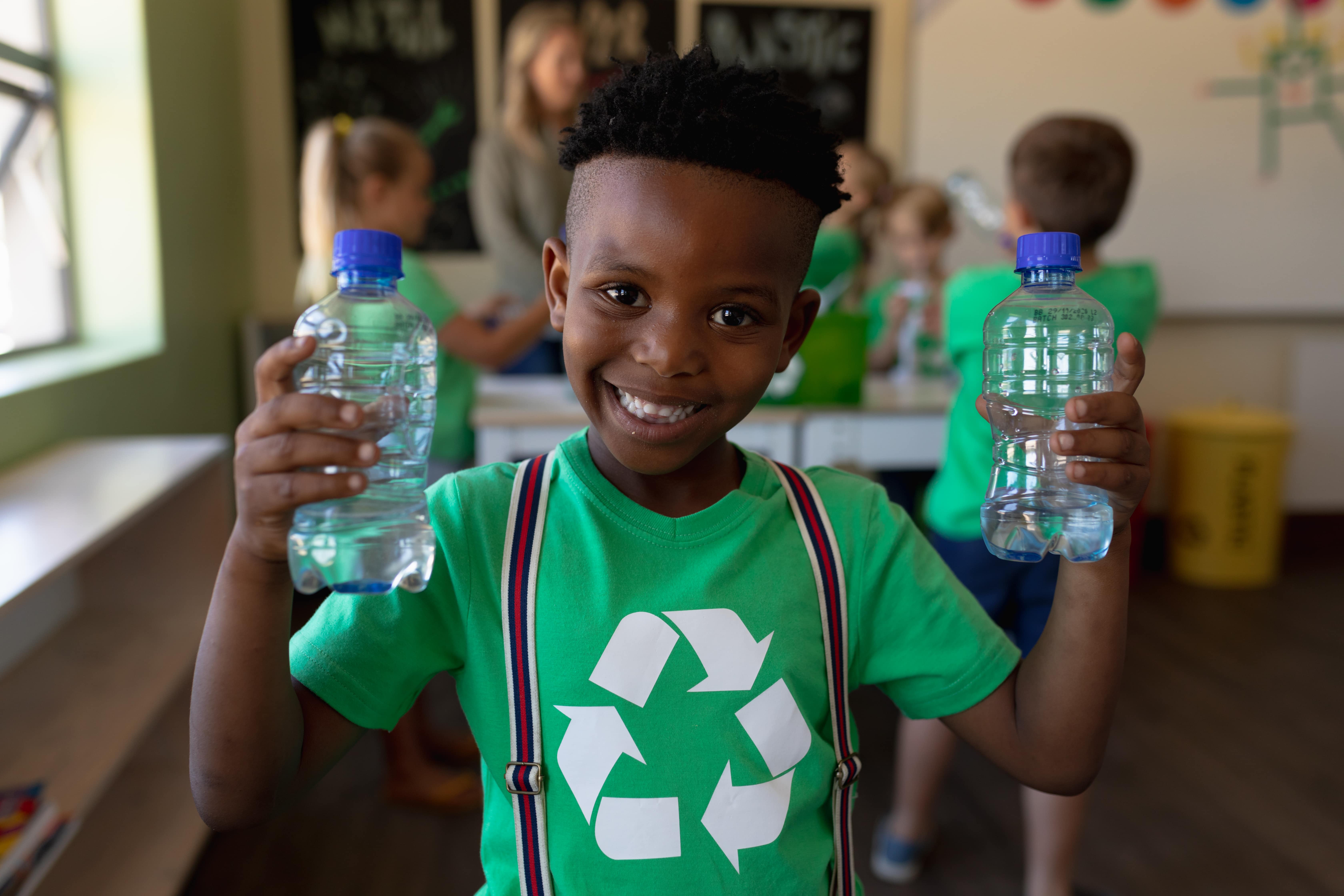 A young boy wearing a green shirt with a recycling symbol on it holding two empty plastic water bottles and learning how to recycle through fun activities for kids.